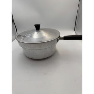 May include: A vintage aluminum saucepan with a lid and a black handle. The pot has a rounded body and a lid with a black knob. The handle is attached to the side of the pot. The pot is designed for cooking.