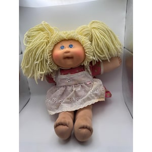 May include: A vintage doll with blonde yarn pigtails, blue eyes, and rosy cheeks. The doll wears a red floral print dress under a white eyelet apron. The doll has tan arms and legs and is a classic toy.