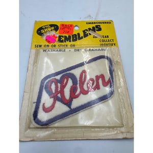 May include: An embroidered patch in a yellow retail card package. The patch is white with a dark blue border and a dark blue heart shape. The name Helen is stitched in red cursive script across the center. The packaging text reads The Show-Offs Emblems.