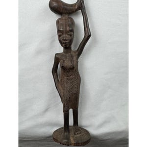 May include: A carved wooden statue of a person carrying an object on their head and holding their arm up. The statue is brown and has a textured surface. The figure is standing on a round base.