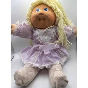 May include: A vintage doll with a soft, round face, blue eyes, and blonde yarn hair. The doll wears a lavender dress with white lace trim and a matching bow. It also has lace-trimmed stockings.