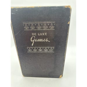 May include: A close-up of a black box with the words "DE LUXE Games" in white script. The box has decorative white floral patterns above and below the text. The edges of the box show signs of wear.