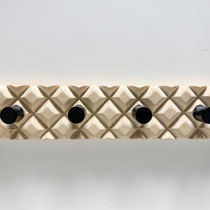 May include: A wooden wall-mounted coat rack with four black pegs. The rack is made of light-colored wood with a geometric, diamond-shaped pattern carved across its surface. The pegs are evenly spaced and cylindrical.