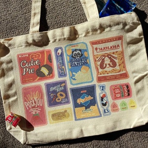May include: A white canvas tote bag with colorful printed boxes featuring various snack foods and the text "Cutie Pie", "Pockyou", "Super Rizz", "Ah-Choo", "Meili Goodbye Panda", "Crackers Hahaha", "Putes Cheeree", and "Aww Flake".