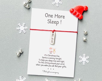 May include: A Christmas-themed gift set on a gray background. It includes a card with the text "One More Sleep!" and a red string bracelet with a "BELIEVE" charm. Decorative elements include snowflakes, bells, and a red Santa hat.