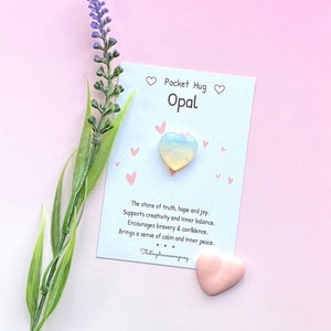 May include: A white card with pink hearts and the text "Pocket Hug Opal" printed on it. A small, heart-shaped opal crystal is on the card. The card also includes text about the meaning of opal, including "The stone of truth, hope and joy." and "Brings a sense of calm and inner peace."