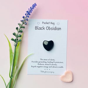 May include: A small black obsidian heart-shaped crystal on a white card with the text "Pocket Hug Black Obsidian" and a description of the stone's properties. The card is on a pink background with a lavender flower.