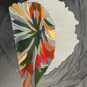 Pode incluir: Um tapete abstrato colorido com fundo cinza. O tapete &eacute; feito de l&atilde; e tem um pelo alto. O tapete tem a forma de um semic&iacute;rculo.