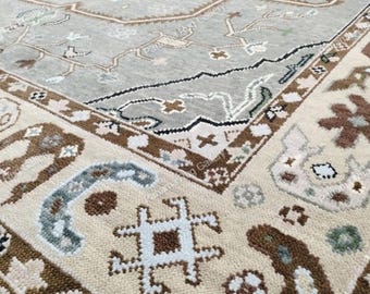 May include: Close-up of a large, intricately patterned rug. The rug features a central field of muted gray with a complex geometric design. The border is a mix of beige, brown, and blue, with repeating floral and abstract motifs. The rug's texture appears soft and inviting.