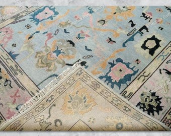 May include: A light blue rug with a floral pattern in pink, orange, and blue. The rug has a beige border with a geometric design and a fringed edge. The rug is partially folded, revealing the underside.