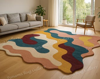 May include: A colorful, abstract area rug with a wavy design in shades of cream, teal, burgundy, yellow, and peach. The rug is placed on a hardwood floor in a living room setting, near a gray sofa and a window.