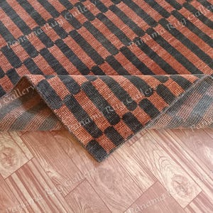 Könnte beinhalten: Ein rechteckiger Teppich mit einem sich wiederholenden Muster aus abwechselnd orangefarbenen und dunkelgrauen Streifen und Blöcken. Der Teppich ist teilweise gefaltet und zeigt die Unterseite. Der Teppich liegt auf einem Holzboden.