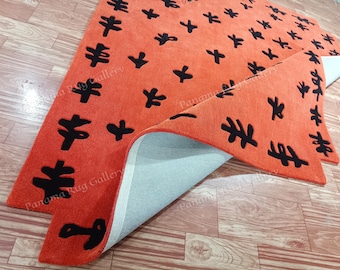 May include: An orange rug with a black abstract pattern. The rug is folded over, revealing a light blue underside. The rug's design features various black shapes, including tree-like and cross-like motifs, creating a modern, artistic look. The rug is placed on a wooden floor.
