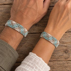 Può includere: Due braccialetti intrecciati abbinati con un motivo testurizzato nei toni del turchese, beige e bianco. I braccialetti sono realizzati con piccoli fili strettamente annodati, creando una fascia uniforme e flessibile attorno al polso.