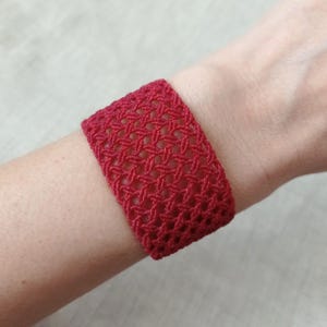 Puede incluir: Una pulsera ancha de ganchillo roja con un patrón geométrico. La pulsera está hecha de un hilo grueso y tiene una apariencia texturizada. El color es un rojo intenso y vibrante. Este accesorio hecho a mano está diseñado para ser usado en la muñeca.