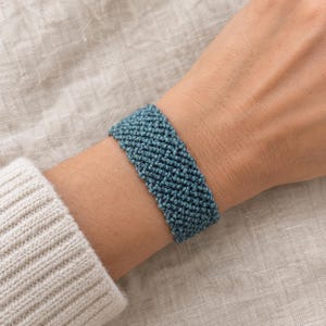 Puede incluir: Una pulsera ancha que presenta un patrón de nudos denso e intrincado en un color azul cerceta profundo. El accesorio está construido con múltiples hilos de cordón tejidos juntos para crear una superficie texturizada y uniforme en la muñeca.