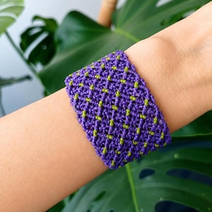 Puede incluir: Una pulsera de macramé ancha que presenta un patrón tejido complejo en cordón morado oscuro con pequeños acentos de hilo verde lima brillante espaciados uniformemente e integrados en el nudo, creando un aspecto texturizado y geométrico.