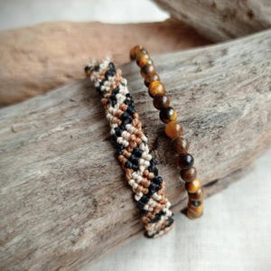 Puede incluir: Dos pulseras presentadas juntas. Una es una pulsera de macramé tejida con un patrón diagonal repetitivo en hilo negro, tostado y crema. La segunda es una pulsera de cuentas compuesta por cuentas redondas, lisas y pulidas de ojo de tigre.
