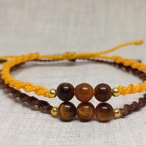 Puede incluir: Dos pulseras de cordón ajustable con una fila central de tres cuentas redondas de piedra marrón, acentuadas por pequeñas esferas de metal dorado. Una pulsera está hecha de cordón amarillo brillante y la otra de cordón marrón oscuro.