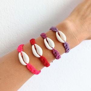 Puede incluir: Cuatro pulseras de amistad coloridas con abalorios de concha blanca. Las pulseras están hechas con un cordón anudado y son en tonos de rosa, rojo y morado.