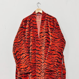 Op de afbeelding: Een kamerjas met lange mouwen en een levendig oranje en zwart tijgerstreeppatroon. Het kledingstuk heeft een sjaalkraag, een bijpassende ceintuur in de taille en een lichtgekleurde voering met dunne verticale strepen bij de halslijn.