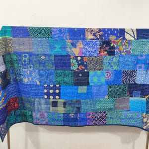 May include: A patchwork quilt in shades of blue, teal, and green. The quilt is made of various fabric squares with different patterns, including floral, geometric, and abstract designs. The quilt is draped over a wooden frame.
