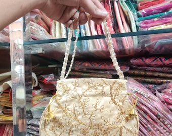 May include: A round, cream-colored drawstring bag with a gold floral embroidery design. The bag has a pearl beaded strap.