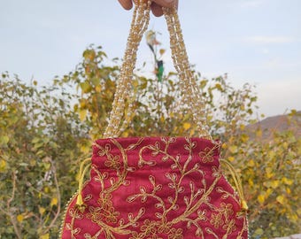 Könnte beinhalten: Eine runde, weinrote Stofftasche mit goldener Stickerei von Blumenmustern. Die Tasche hat einen Kordelverschluss und einen Perl- und Goldkettenriemen.