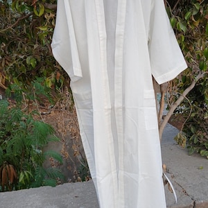 Pode incluir: Uma bata de algod&atilde;o branca com uma frente longa e aberta e um cinto para amarrar. A bata tem mangas compridas e um bolso do lado esquerdo.