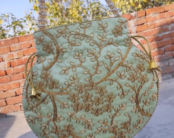 May include: A light green, round handbag with gold floral embroidery. The bag has a drawstring closure and a beaded strap. Gold tassels hang from the bottom. The bag is held up against a brick wall and a blue sky.