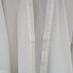 Pode incluir: Uma bata de algod&atilde;o branca de manga comprida com um cinto para amarrar. A bata tem um design simples e &eacute; feita de um tecido leve.