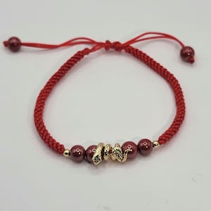 May include: A red string bracelet with a gold snake charm and burgundy beads.
