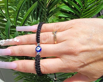 May include: Black woven bracelet with a blue and white evil eye bead. The bracelet is adjustable and has a silver-colored bead at the end. A gold ring with a blue stone is also visible.