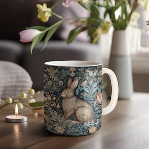 May include: A white ceramic mug with a blue and green floral pattern featuring a brown rabbit. The rabbit is sitting in the center of the mug, surrounded by flowers and leaves.