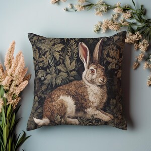 May include: Square decorative pillow featuring a detailed illustration of a brown and white rabbit with long ears, set against a dark background of stylized foliage. The pillow has a soft, textured appearance, with a light blue background.