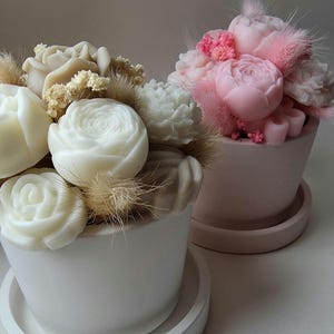 Könnte beinhalten: Zwei kleine Keramiktöpfe, einer weiß und einer rosa, gefüllt mit Seifenblumenarrangements. Der weiße Topf hat weiße Seifenrosen und getrocknete Gräser, der rosa Topf rosa Pfingstrosenseifen und Akzente. Beide stehen auf Untertassen.