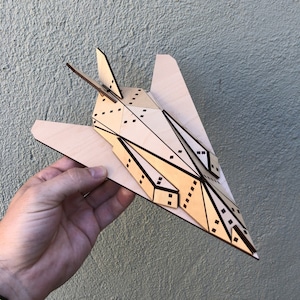 May include: A wooden model of a fighter jet with black lines and details. The model is made of light brown wood and is held in a hand.