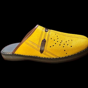 May include: A yellow leather clog-style slipper with a brown trim and accents. The shoe features decorative stitching, a series of small perforations, and a dark brown strap. The sole is a combination of gray, brown, and black.