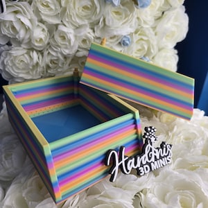 May include: A small, open box with a lid, featuring a vibrant rainbow striped design. The box has a light blue interior and a yellow frame. The lid is placed next to the box. The logo "Handonis 3D MINIS" is visible.