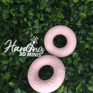 May include: Two light pink circular rings featuring embossed daisy flower patterns and the text "HandAnis 3D MINIS" on the surface. The objects have a smooth, matte finish and a central hole, presented as a pair of decorative items.