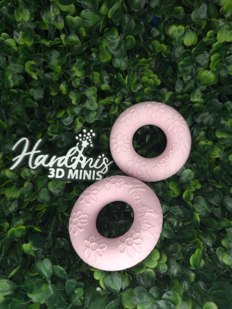 May include: Two light pink circular rings featuring embossed daisy flower patterns and the text "HandAnis 3D MINIS" on the surface. The objects have a smooth, matte finish and a central hole, presented as a pair of decorative items.