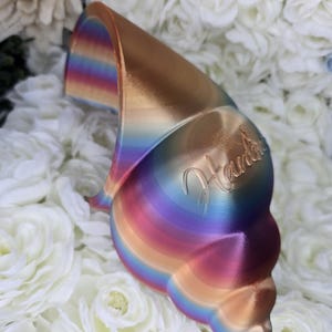 May include: A seashell-shaped decorative object with a metallic, iridescent finish. The colors shift from gold to blue, purple, and pink. The word "Handmade" is inscribed on the surface. The object is resting on a bed of white flowers.