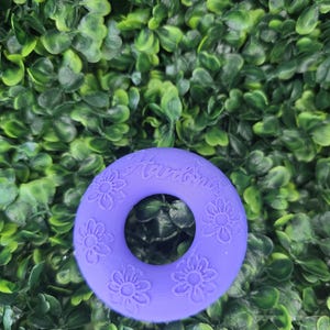 May include: A circular purple silicone ring featuring embossed flower patterns and the text "Hardines" engraved on the top surface. The object has a smooth, matte finish and a central hole, displaying a consistent violet color throughout the design.