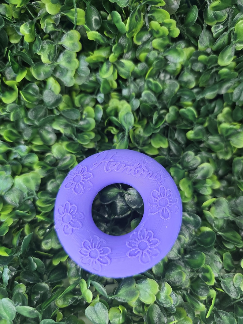 May include: A circular purple silicone ring featuring embossed flower patterns and the text "Hardines" engraved on the top surface. The object has a smooth, matte finish and a central hole, displaying a consistent violet color throughout the design.