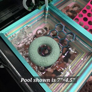 May include: A rectangular teal pool measuring 7 inches by 4.5 inches, filled with water and smooth pebbles. A light green donut-shaped float with embossed flower patterns and a dark blue honeycomb-shaped accessory float on the water surface.