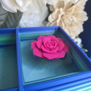 May include: A bright pink 3D printed rose flower sits centered inside a square blue container. The container features a gradient of blue and green horizontal stripes along its outer walls and a smooth, light blue base surface inside the box.