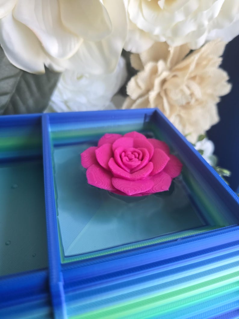 May include: A bright pink 3D printed rose flower sits centered inside a square blue container. The container features a gradient of blue and green horizontal stripes along its outer walls and a smooth, light blue base surface inside the box.