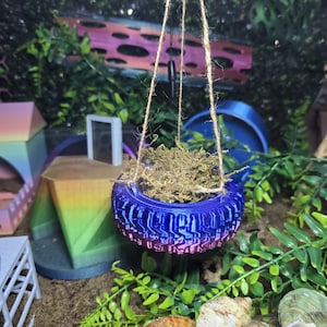 May include: A hanging planter shaped like a miniature tire with a gradient blue and purple finish, suspended by three strands of natural jute twine. The tire contains a small amount of dried moss and is designed for small animal enclosures.