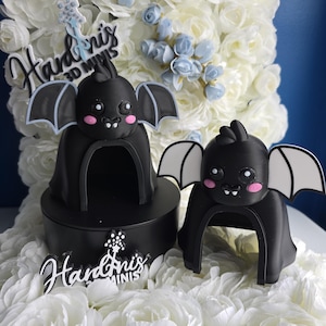 May include: Two black bat-shaped figurines with pink cheeks and white-edged wings. One bat sits atop a black circular base. The figurines are surrounded by white and light blue floral decorations. The text "Handoris 3D Minis" is visible.