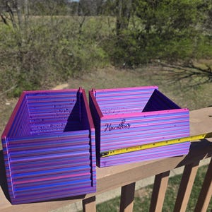 May include: Two rectangular storage bins with a horizontal striped pattern in shades of pink, purple, and blue. A yellow measuring tape is placed across the front of the right bin, showing a width of approximately 5 inches for the container.